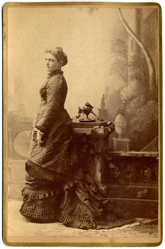 Cabinet Card Photograph full length portrait of Madelon Stockwell Turner as a young woman. She is wearing a dark ruffled dress and standing in front of a garden background. 
