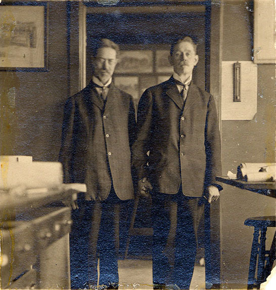 The brothers are standing between a table and set of drawers covered with large rolls of paper. They are dressed in suits and holding hands.