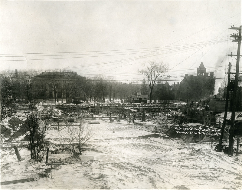 Snow covers wooden scaffolding and bare ground of construction site. Workers carry lumber and climb scaffolding; Alumni Memorial Hall and Newberry Hall are visible in the distance.