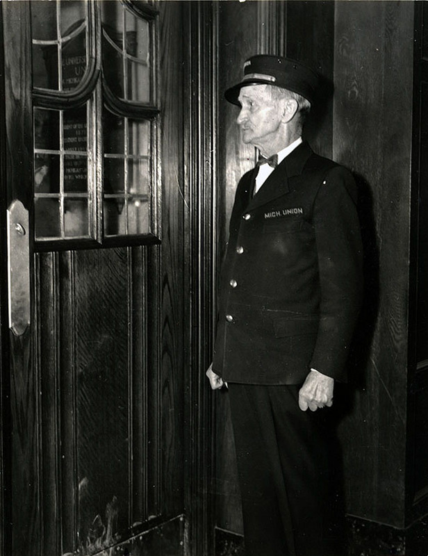 Photograph. Man in doorman's uniform and cap stands by door, arms by his sides and fists balled. He is staring intently out the window.