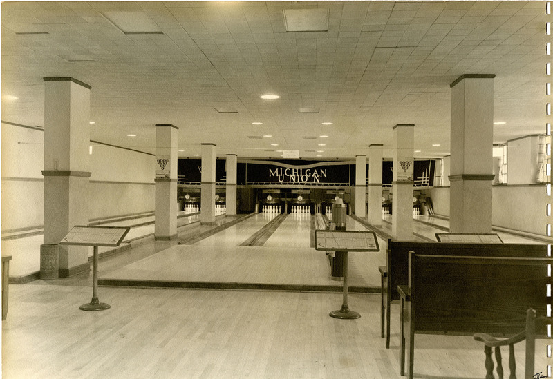 Photograph. Seven-lane bowling alley; empty, with pins set and scorecards on stands.