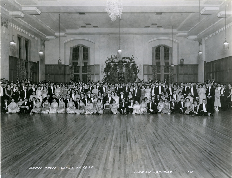 Large, coed group of students in formal dress gather at one end of ballroom for group photo. The first two rows are seated on the floor and the rest standing behind.
