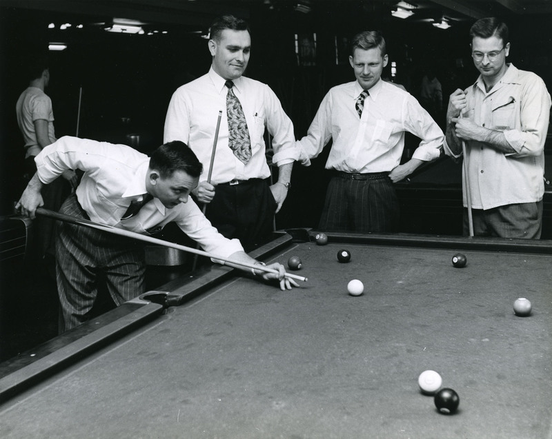 Four young men with pool cues and rolled-up shirtsleeves gather around a billiards table. One prepares to take a shot.