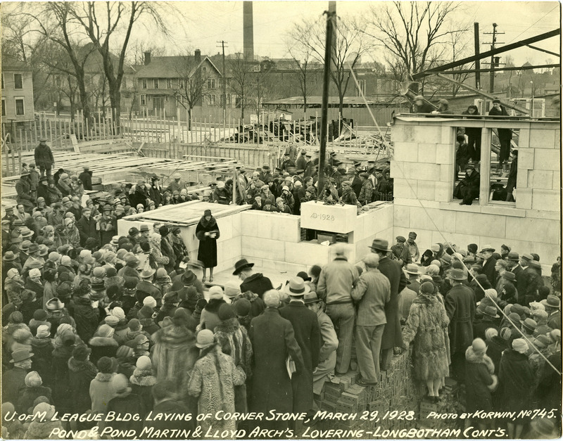 Cornerstone inscribed "AD 1928" being lowered into place as a woman reads from a piece of paper. Crowd of spectators wear winter hats and coats and stand on or near unfinished walls.